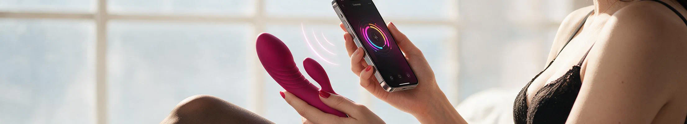Eine Frau in schwarzer Unterwäsche hält ein app-gesteuertes Sextoy in der einen und ein Smartphone in der anderen Hand.