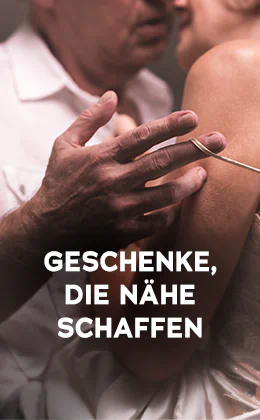 Das Bild zeigt eine Werbeanzeige, wo eine Frau und ein Mann dicht aneinander stehen. Der Mann trägt ein weisses Hemd und die Frau ein helles Oberteil mit dünnen Trägern. Die Köpfe sind dicht beieinander und der Mann macht mit seiner Hand den dünnen Träger der Frau an ihrer Schulter runter. Plakativ steht in großer, weisser Schrift "Geschenke, die Nähe schaffen". Unten befindet sich ein rotfarbener Button mit der weissen Aufschrift „Zeig’s mir“. Der Hintergrund ist dunkel gehalten.