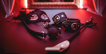 Auf einem dunklen Tisch liegen BDSM-Accessoires: schwarze Augenmaske mit Spitze, Ballgag, Lederfesseln, Reitgerte und weitere Utensilien. Beleuchtung in Rot schafft eine erotische Atmosphäre.